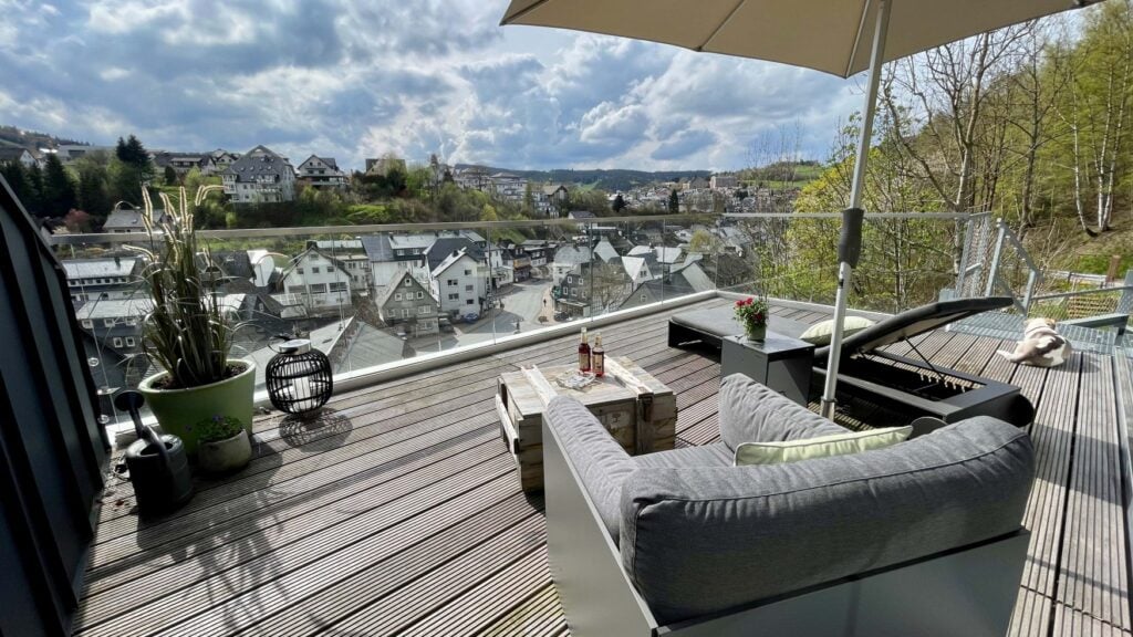 Eine Dachterrasse mit Holzdielen; ein Polstersessel und ein Liegestuhl stehen unter einme Sonnenschirm; rechts liegt ein Hund, mittig eröffnet sich das Panorama auf Willingen im Sauerland