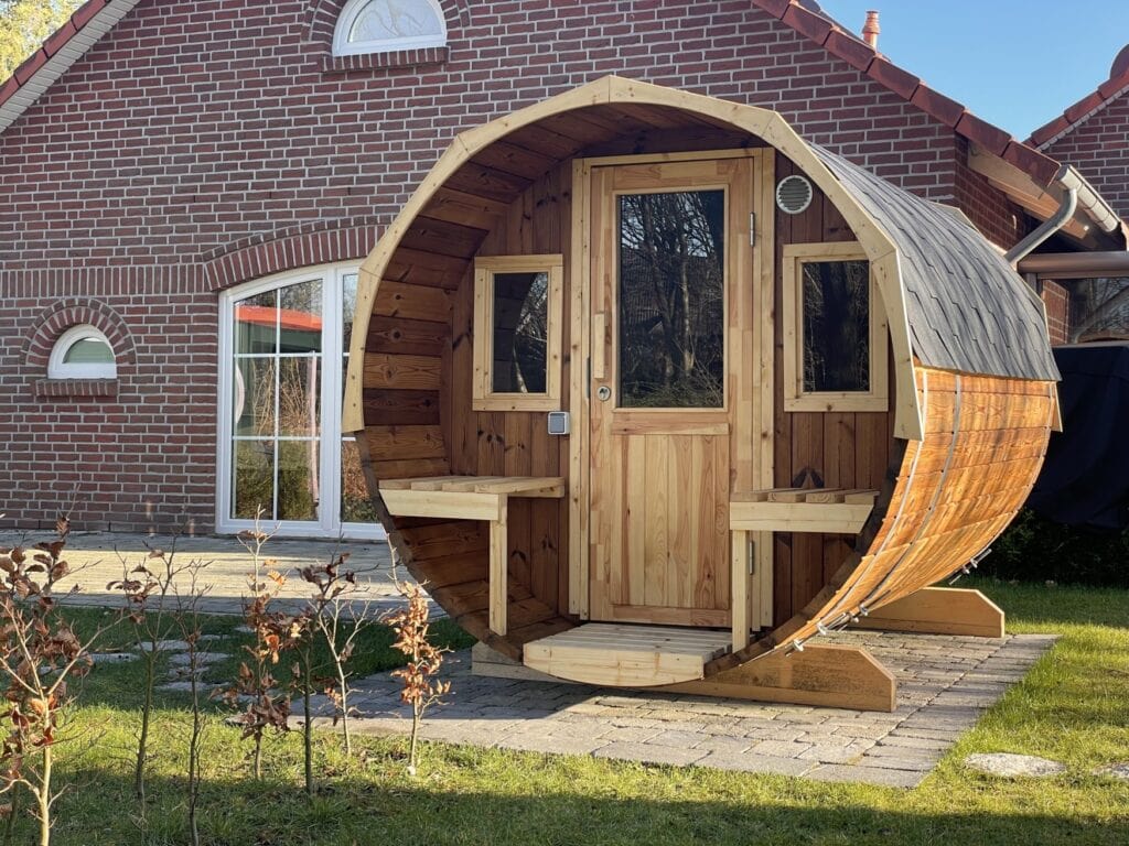 Vor einem rotgeziegelten Haus steht ein großes Saunaholzfass auf einer Wiese