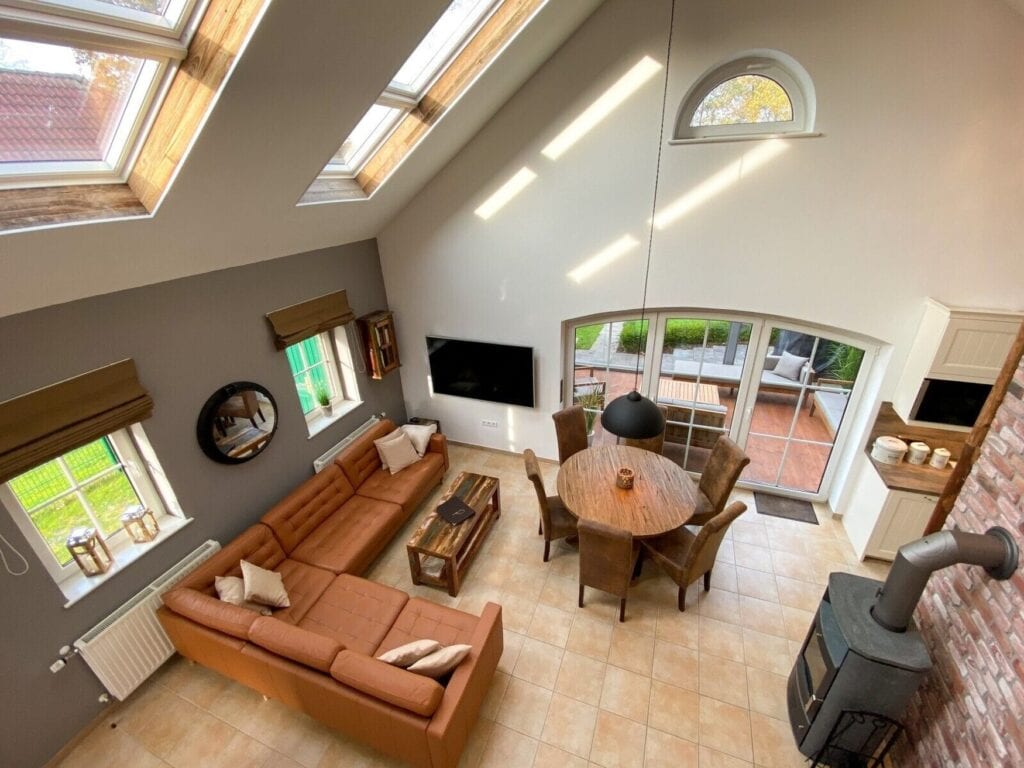 Ein Wohnzimmer mit Decke bis in den 1. Stock von oben gesehen; oben zwei Dachfenster, links ein braunes Lederecksofa, mittig ein runder Tisch mit sechs Stühlen, rechts ein freistehender Metallofen an einer Ziegelwand
