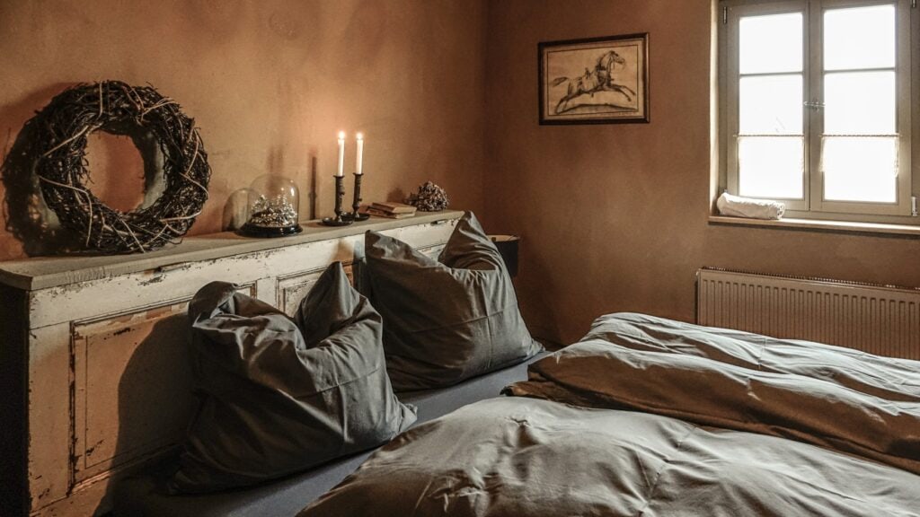 Ein Bett mit zwei Sätzen Bettwäsche, dahinter ein rustikales Holzkopfteil mit zwei Kerzen und einem Holzflechtkranz; rechts im Bild ein Fenster