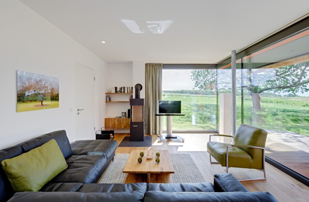 Ein Wohnzimmer; links und mittig im Vordergrund eine schwarze Ledereckcouch und ein hölzerner Couchtisch, rehts davon ein grüner Ledersessel mit Metallrahmen; im Hintergrund ein moderner, freistehender Kamin und ein freistehender Fernseher. Die rechte und hintere Wand des Raums besteht aus bodentiefen Fenstern, die den Blick auf einen Rasen freigeben