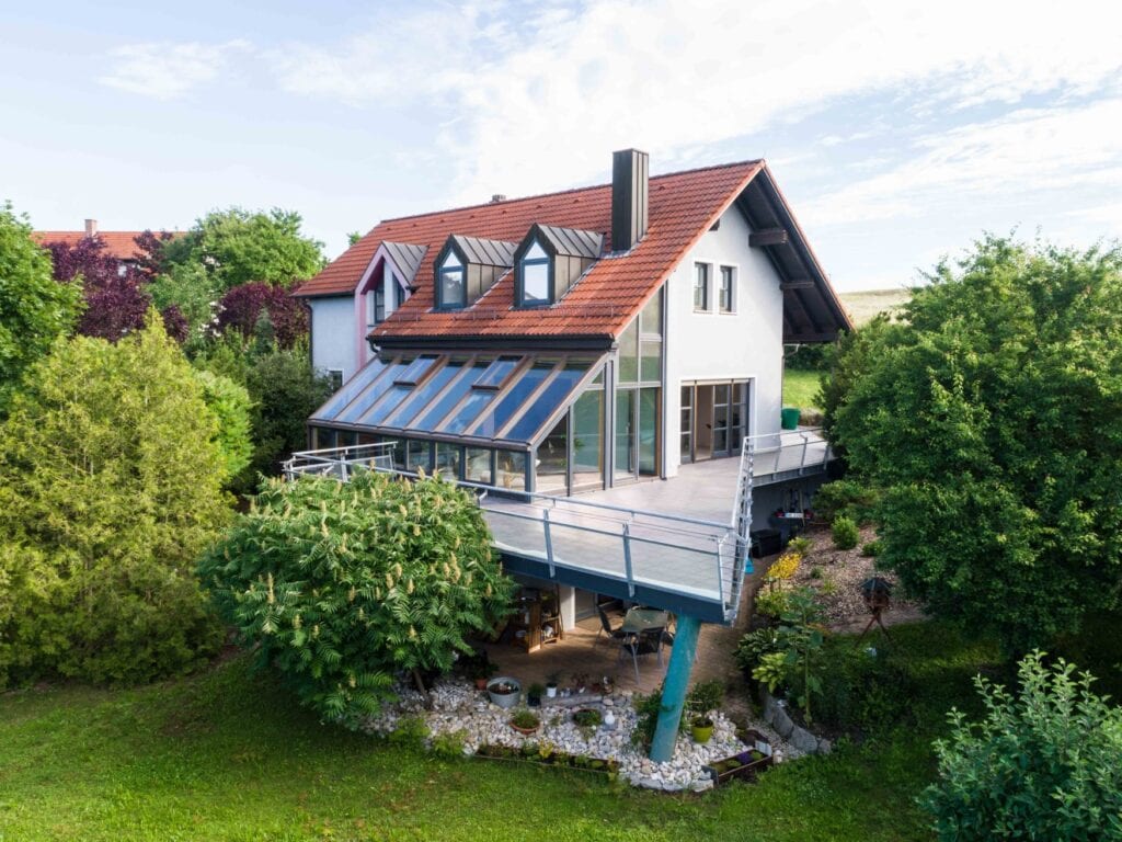 Ein Haus mit großem Balkon-Wintergarten, rotem Dach und Garten mit vielen Bäumen