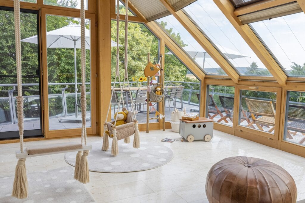 Ein Wintergarten mit hellen Bodenfliesen, einer Schaukel, einem runden Lederhocker; durch eine Tür geht es auf einen Balkon, dahinter sind Bäume zu sehen