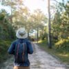 Ein Mann mit Rucksack und Anglerhut spaziert durch einen Wald; man sieht ihn von hinten