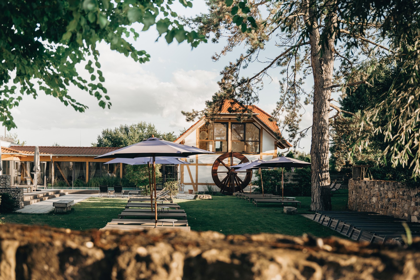 In the middle of a lawn stands a row of sun loungers with parasols. Behind them rises a modern half-timbered building with a watermill wheel on the façade.