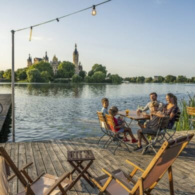 Auf einem hölzernen Steg vor einem Gewässer stehen mehrere Liegestühle und kleine Holztische; links zwei Fahrräder, rechts eine Familie mit drei Kindern und Getränken an einem der Tische. Auf der anderen Seite des Sees erhebt sich gut sichtbar das Schweriner Schloss im Abendlicht