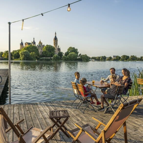 Auf einem hölzernen Steg vor einem Gewässer stehen mehrere Liegestühle und kleine Holztische; links zwei Fahrräder, rechts eine Familie mit drei Kindern und Getränken an einem der Tische. Auf der anderen Seite des Sees erhebt sich gut sichtbar das Schweriner Schloss im Abendlicht
