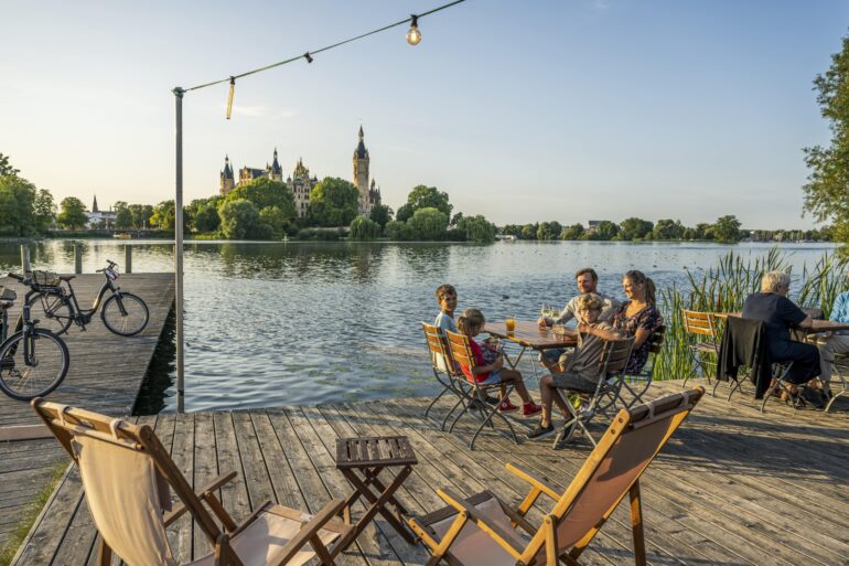 Auf einem hölzernen Steg vor einem Gewässer stehen mehrere Liegestühle und kleine Holztische; links zwei Fahrräder, rechts eine Familie mit drei Kindern und Getränken an einem der Tische. Auf der anderen Seite des Sees erhebt sich gut sichtbar das Schweriner Schloss im Abendlicht