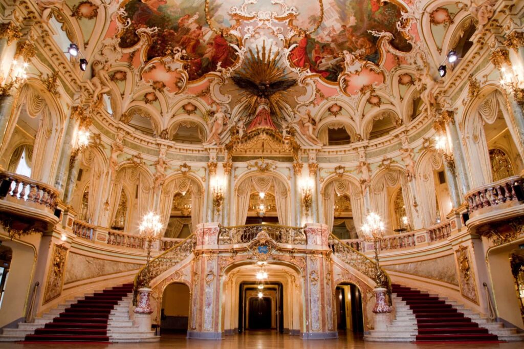 The foyer in the main building of the Wiesbaden State Theater, a round hall decorated with frescoes and stucco. Two grand staircases lead up to a gallery.