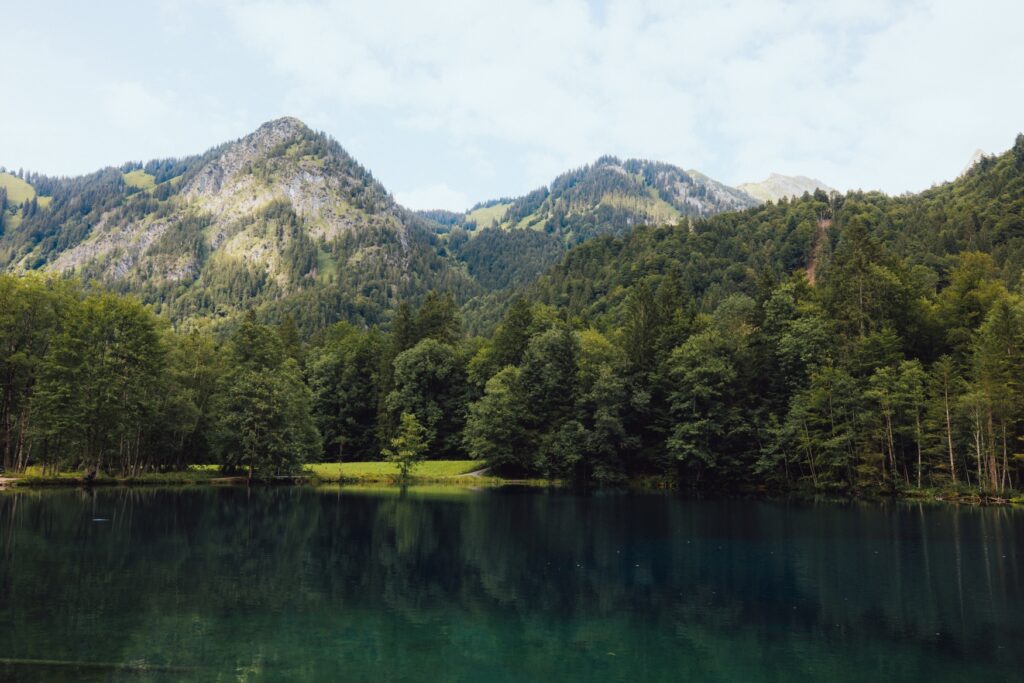 Ein blau-grüner Bergsee, der sich von links nach rechts durchs Bild zieht. Dahinter ein dicht bewaldetes Ufer, hinter dem zwei hohe Berge auftauchen, unter blauem Himmel