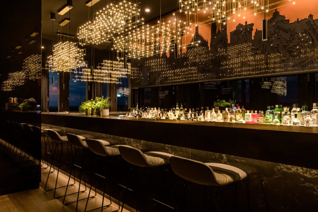 Eine lange Hotelbar; vor der Bar stehen fünf Polsterhocker mit sehr niedriger Lehne. Die Bar selbst ist in Marmor und dunklem Metall gehalten. Über dem Tresen hängen mehrere Leuchten, die aus gold lackiertem Draht und zahlreichen kleinen Lampen bestehen. Hinter der Bar befindet sich eine Vielzahl unterschiedlicher Flaschen.
