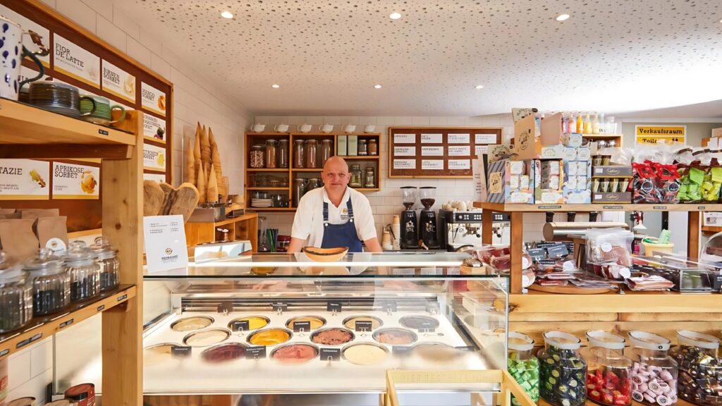 The sales area of an ice cream parlor. Behind the counter stands a bald man wearing a blue apron, smiling warmly. There are 14 flavors of ice cream to choose from on the counter. To the right of the ice cream counter is another shelf with specialties ranging from tea to Black Forest ham to individually packaged hand soaps. The interior is decorated in light wood.