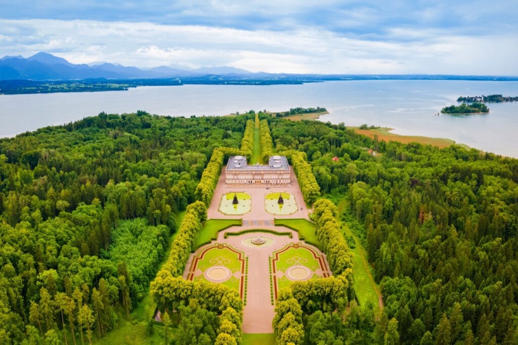Aerial shot of a castle with a long park in front and behind of it. To the left and right of the park is a thick forest, the whole area is on an half-island inside of a lake. Far in the background on the other side of the lake lay the alps.