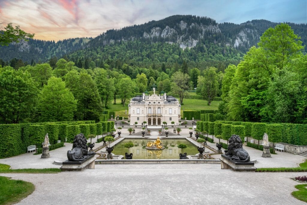 A small, white palaca building in a symmetric, accurate garden. In front of the main entrance is a rectangular pool with a golden statue in the middle. Left and right are some rectangular shrubberies. Behind the palace lays a mountain, it's slope covered by a dense forest.