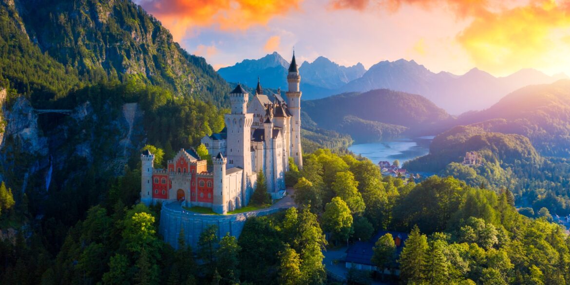 The Neuschwanstein Castle, with it's red front and it's many towers, perched on the slope of a mountain in the Alps. In the background is a valley with a lake and a small town, all around is a lush, green forest. Everything is illuminated by a golden and orange dawn.