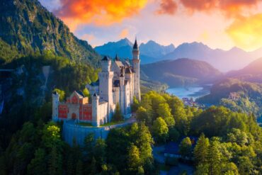 The Neuschwanstein Castle, with it's red front and it's many towers, perched on the slope of a mountain in the Alps. In the background is a valley with a lake and a small town, all around is a lush, green forest. Everything is illuminated by a golden and orange dawn.