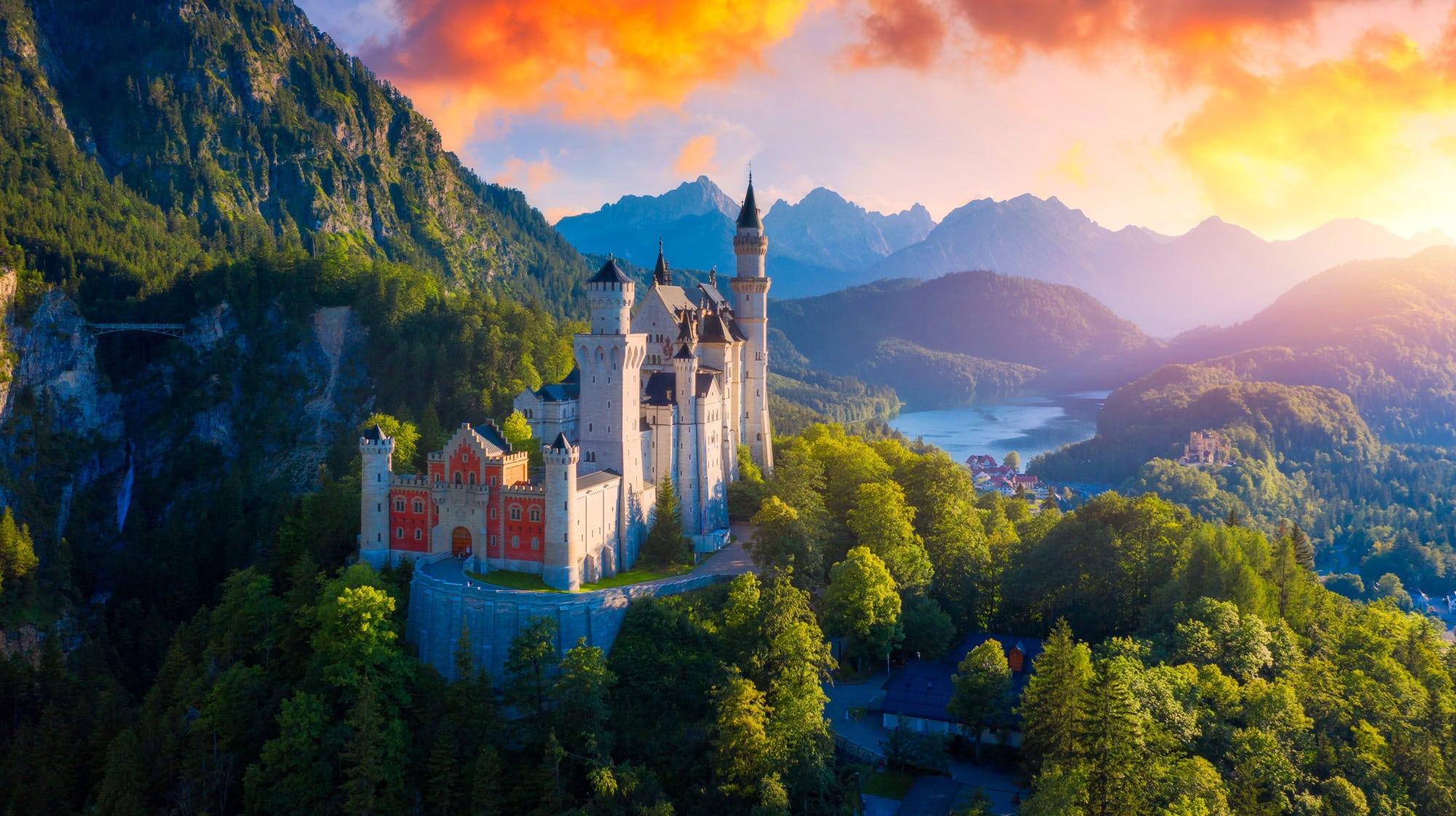 The Neuschwanstein Castle, with it's red front and it's many towers, perched on the slope of a mountain in the Alps. In the background is a valley with a lake and a small town, all around is a lush, green forest. Everything is illuminated by a golden and orange dawn.