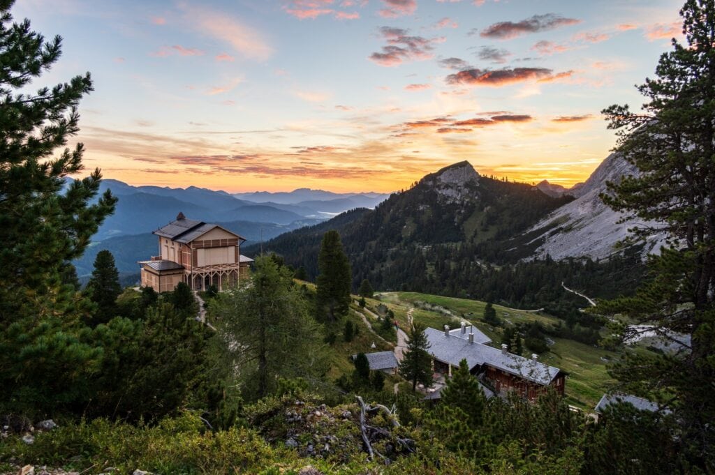 Two houses on a cliff in the Alps; on the right there is only a roof visible with the rest of the house covered by trees and bushes. On the right is a wooden house with two stories. It looks like a typical alpine hut, it's front towards the valley. To the right of the cliff, a stoney mountain rises to the sky which is coloured a bright yellow by the setting sun