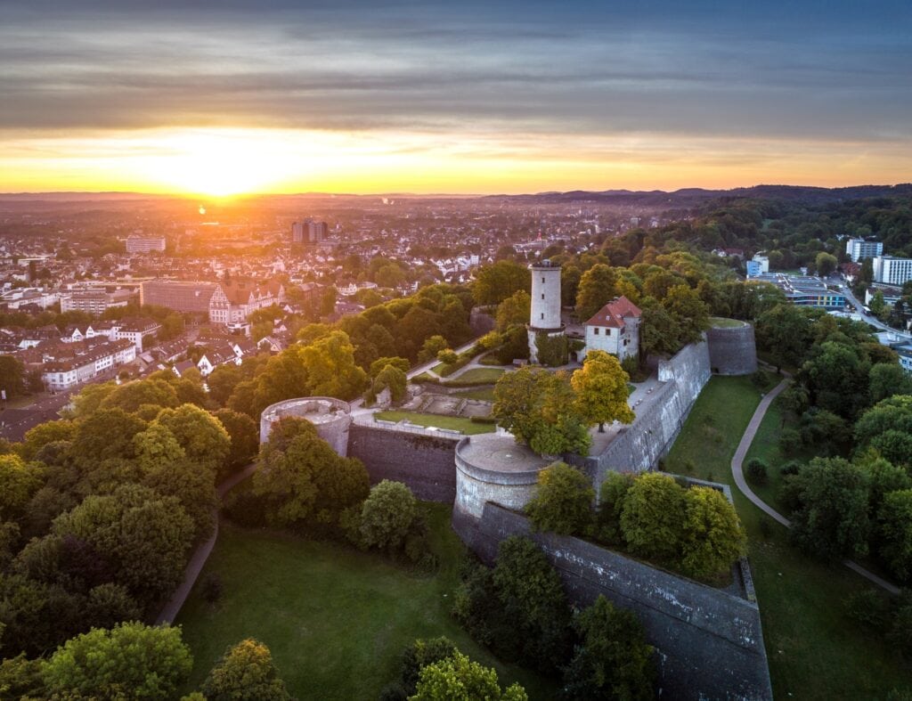 Eine mächtige Burganlage mit den Fundamenten von vier Rundtürmen an den Ecken aus der Vogelperspektive. Im Burghof stehen noch ein Wohngebäude und ein Rundturm, der Berg rund um die Burg ist bewaldet. Links und rechts des Berges erstreckt sich die Stadt Bielefeld. Ein Sonnenuntergang am Horizont taucht alles in ein goldenes Licht