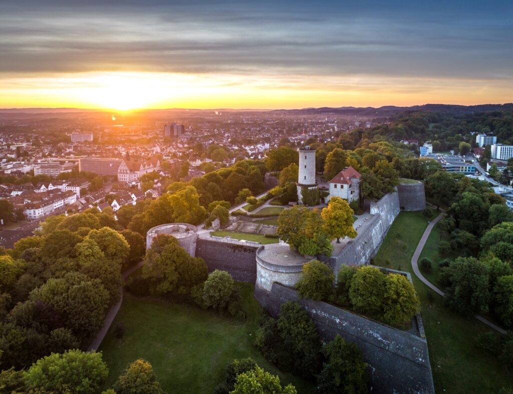 A mighty castle complex with the foundations of four round towers at the corners, seen from a bird's eye view. In the castle courtyard, there is still a residential building and a round tower, and the hill surrounding the castle is wooded. To the left and right of the hill lies the city of Bielefeld. A sunset bathes everything in a golden light.
