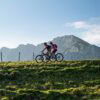 Zwei Mountainbiker fahren auf einem Hügelkamm von rechts nach links durhcs Bild. Im Hintergrund erheben sich unter blauem Himmel mehrere Bergspitzen