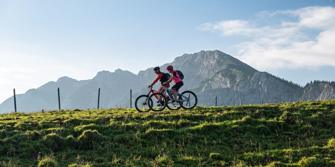 Zwei Mountainbiker fahren auf einem Hügelkamm von rechts nach links durhcs Bild. Im Hintergrund erheben sich unter blauem Himmel mehrere Bergspitzen
