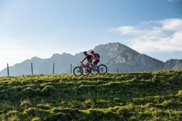Zwei Mountainbiker fahren auf einem Hügelkamm von rechts nach links durhcs Bild. Im Hintergrund erheben sich unter blauem Himmel mehrere Bergspitzen