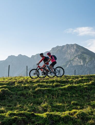 Zwei Mountainbiker fahren auf einem Hügelkamm von rechts nach links durhcs Bild. Im Hintergrund erheben sich unter blauem Himmel mehrere Bergspitzen