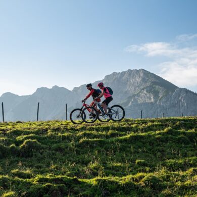 Zwei Mountainbiker fahren auf einem Hügelkamm von rechts nach links durhcs Bild. Im Hintergrund erheben sich unter blauem Himmel mehrere Bergspitzen