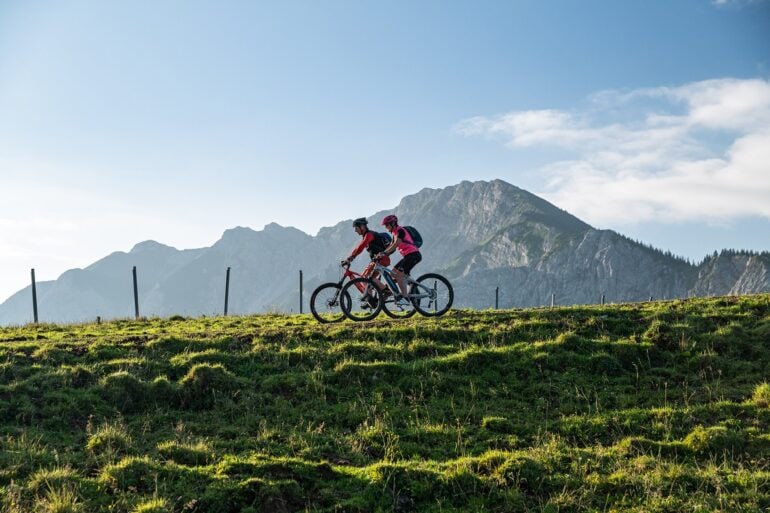 Zwei Mountainbiker fahren auf einem Hügelkamm von rechts nach links durhcs Bild. Im Hintergrund erheben sich unter blauem Himmel mehrere Bergspitzen