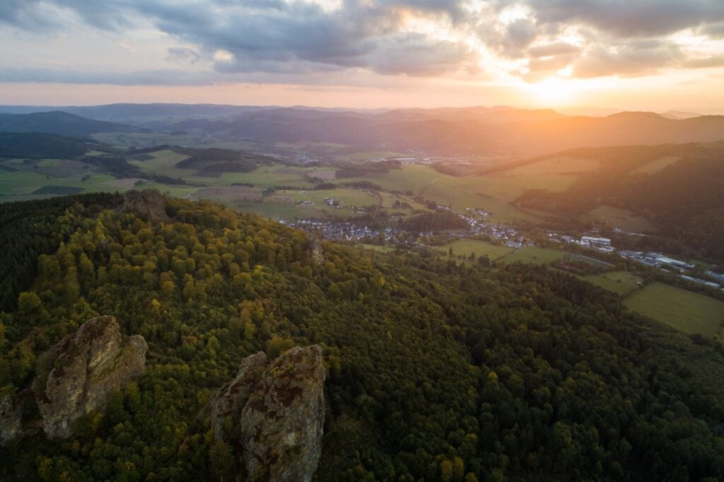 Drohnenaufnahme einer Landschaft im Sauerland; die untere Hälfte des Bildes wird von einem bewaldeten Berg eingenommen, auf dessen Gipfel vier mächtige Felsen aus den Bäumen "emporsteigen". Am Fuße des Berges reihen sich hügelige Felder mit einigen Siedlungen dazwischen aneinander. Am Horizont weitere Hügel, hinter denen die Sonne zwischen einigen Wolken untergeht