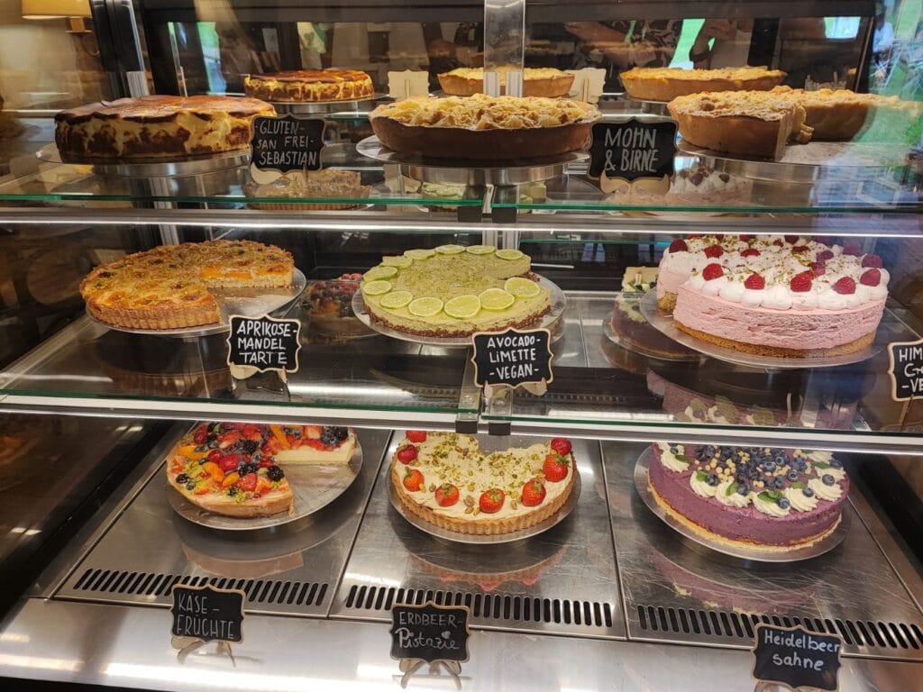 In der gläsernen Auslage eines Cafés stehen in drei Stockwerken jeweils drei Torten, die alle mit einem handgeschriebenen Schild gekennzeichnet sind: Glutenfrei San Sebastian, Mohn & Birne, Aprikose-Mandel, Avocado-Limette (vegan), Käse-Früchte, Erdbeer-Pistazie, Heidelbeer-Sahne; die Schilder der jeweils rechten Torten sind nicht im Bildausschnitt