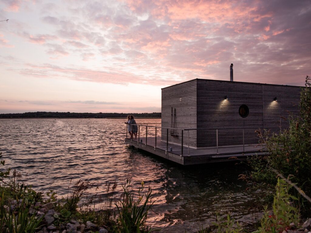 A large lake bathed in a pink sunset; on the right side of the picture, a wooden houseboat floats on the water, with two people leaning against the railing on the deck, looking out at the lake.