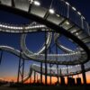 Die Haldenskultpur "Tiger & Turtle" in Duisburg vor der fast untergegangen Sonne; die auf Stelzen befestigte, wie eine Achterbahn verdrehte Treppe ist von vielen kleinen Lampen erleuchtet. Unter der Skulptur stehen einige Menschen