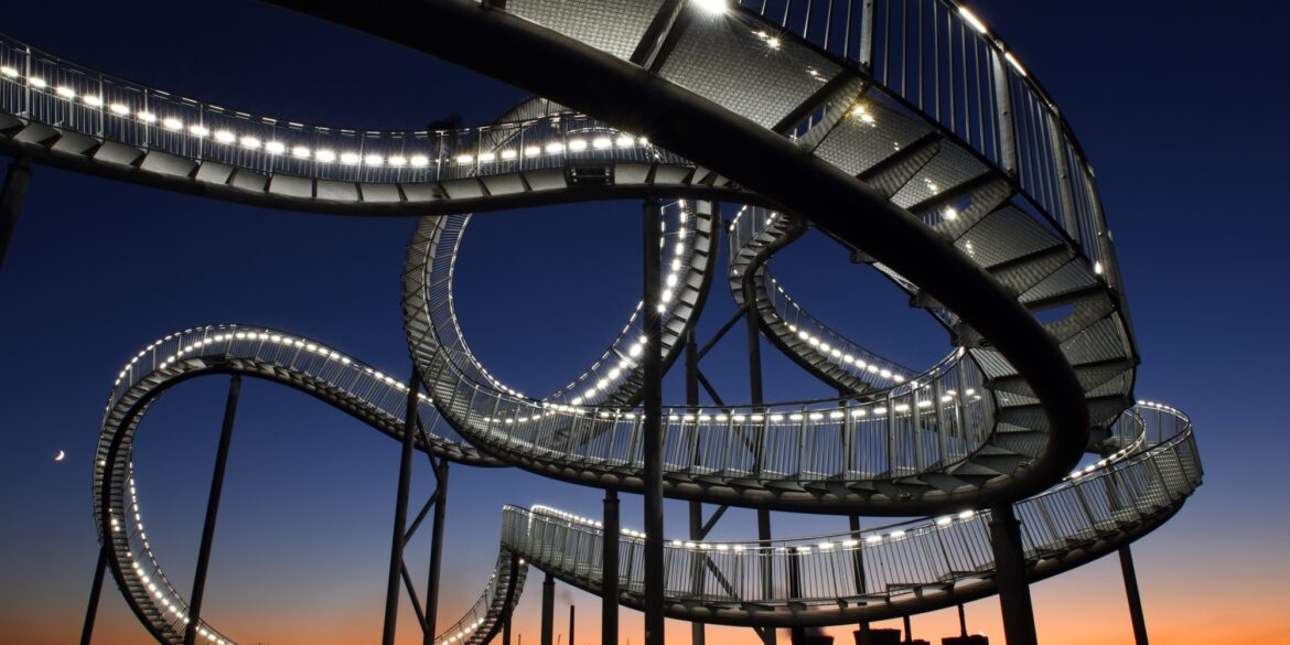 Die Haldenskultpur "Tiger & Turtle" in Duisburg vor der fast untergegangen Sonne; die auf Stelzen befestigte, wie eine Achterbahn verdrehte Treppe ist von vielen kleinen Lampen erleuchtet. Unter der Skulptur stehen einige Menschen