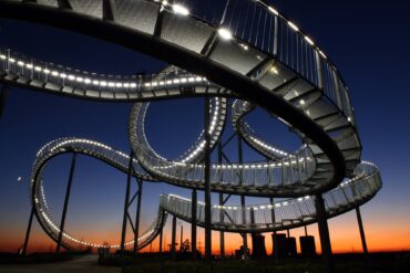 Die Haldenskultpur "Tiger & Turtle" in Duisburg vor der fast untergegangen Sonne; die auf Stelzen befestigte, wie eine Achterbahn verdrehte Treppe ist von vielen kleinen Lampen erleuchtet. Unter der Skulptur stehen einige Menschen