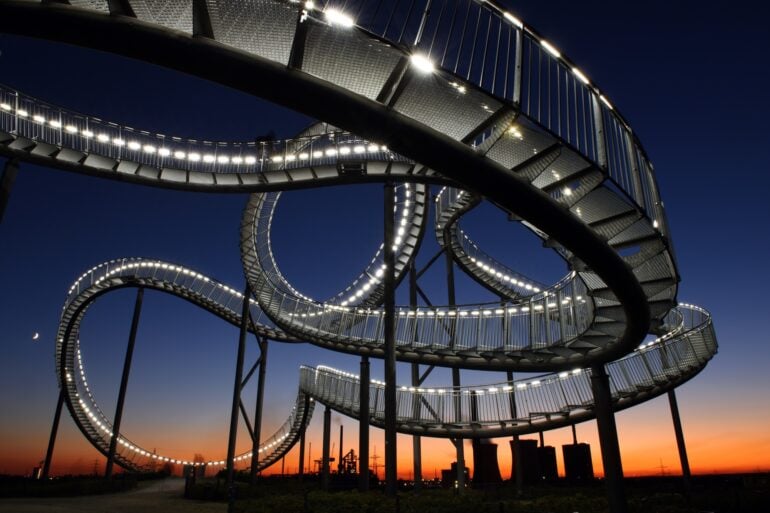Die Haldenskultpur "Tiger & Turtle" in Duisburg vor der fast untergegangen Sonne; die auf Stelzen befestigte, wie eine Achterbahn verdrehte Treppe ist von vielen kleinen Lampen erleuchtet. Unter der Skulptur stehen einige Menschen