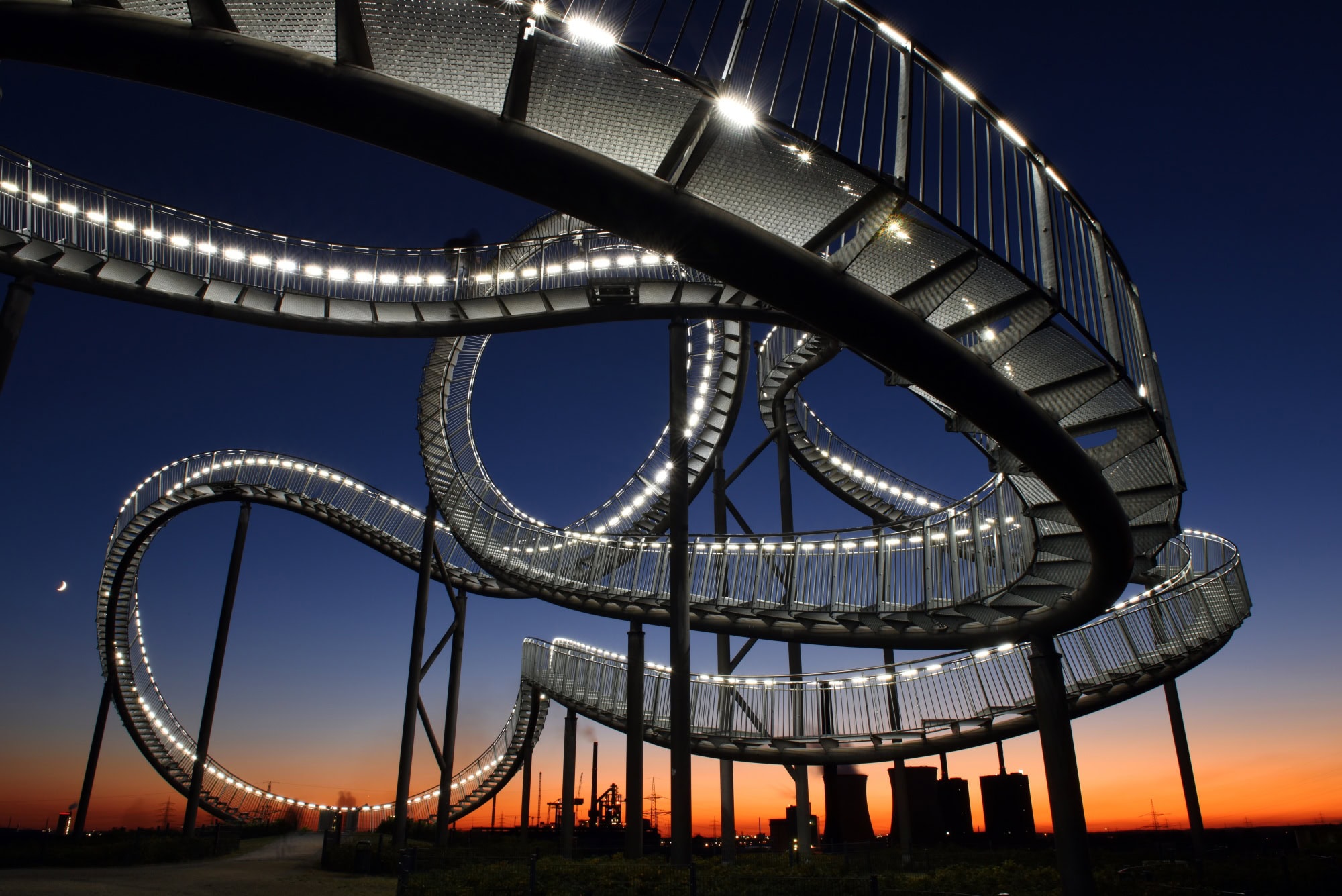 Die Haldenskultpur "Tiger & Turtle" in Duisburg vor der fast untergegangen Sonne; die auf Stelzen befestigte, wie eine Achterbahn verdrehte Treppe ist von vielen kleinen Lampen erleuchtet. Unter der Skulptur stehen einige Menschen
