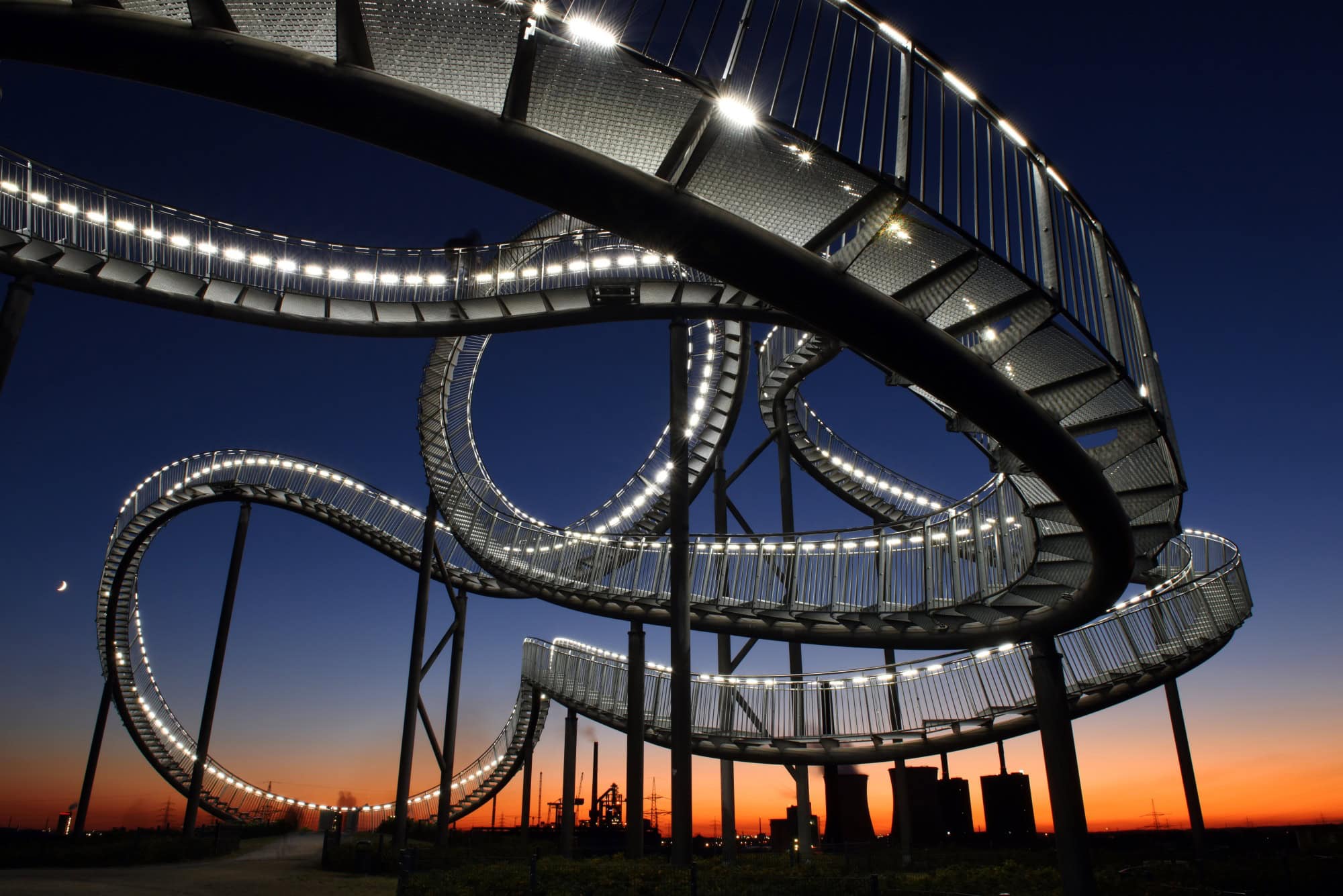 The “Tiger & Turtle” sculpture in Duisburg in front of the almost setting sun; the staircase, mounted on stilts and twisted like a roller coaster, is illuminated by many small lamps. Several people are standing beneath the sculpture.