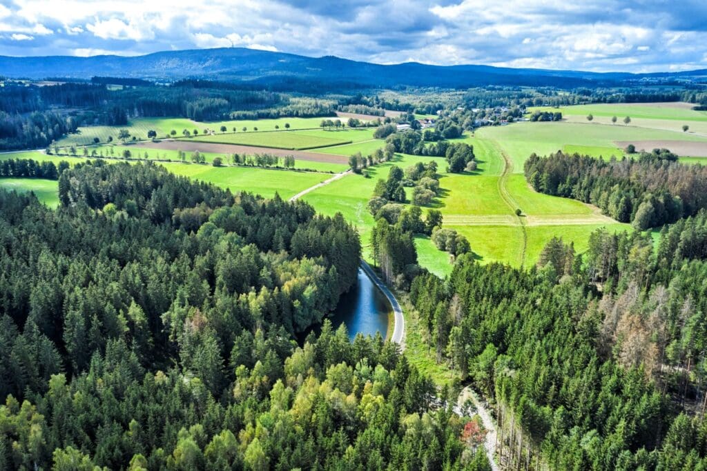 Vogelperspektive auf ein Mittelgebirge. Im Vordergrund ein dichter Nadelwald, in dessen Mitte ein Fluss (die Eger) zu erkennen ist; daneben läuft ein frisch asphaltierter Radweg. Hinter dem Waldstück erstrecken sich Weiden und vereinzelte Bäume, weit im Hintergrund erheben sich die bewaldeten Hänge des Fichtelgebirges