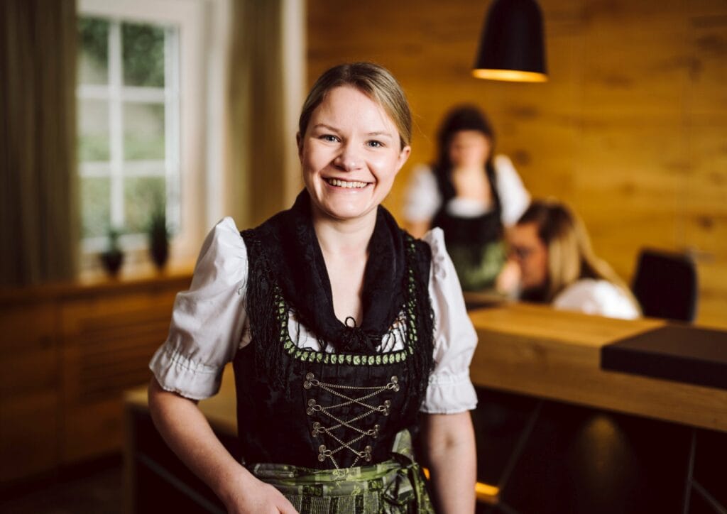 Eine junge Frau mit blondem, zurückgebundenen Haar in einer schwarz-weiß-grünen Schwarzwaldtracht lächelt direkt in die Kamera; hinter ihr sind unscharf zwei weitere Frauen in ähnlicher Tracht an einer Hotelrezeption erkennbar, die Wände sind holzverkleidet