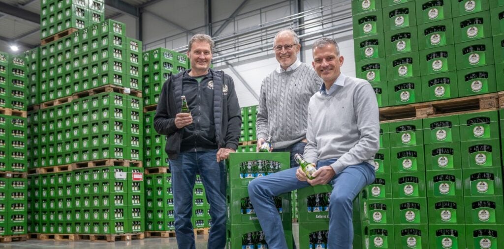 Drei Männer mittleren Alters stehen in einer Lagerhalle. Hinter ihnen stehen bis an die Decke gestapelt zahlreiche Paletten mit grünen Bierkästen. Einer der Männer sitzt auf zwei gestapelten Kästen, hinter ihm stehen die beiden anderen. Alle halten eine Bierflasche in Händen