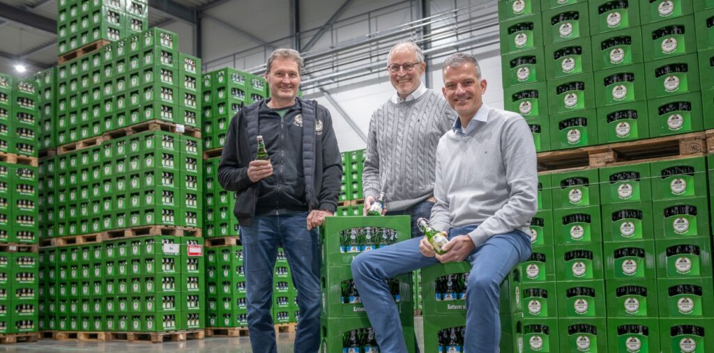 Three middle-aged men are standing in a warehouse. Behind them are numerous pallets stacked up to the ceiling with green beer crates. One of the men is sitting on two stacked crates, with the other two standing behind him. They are all holding beer bottles in their hands.