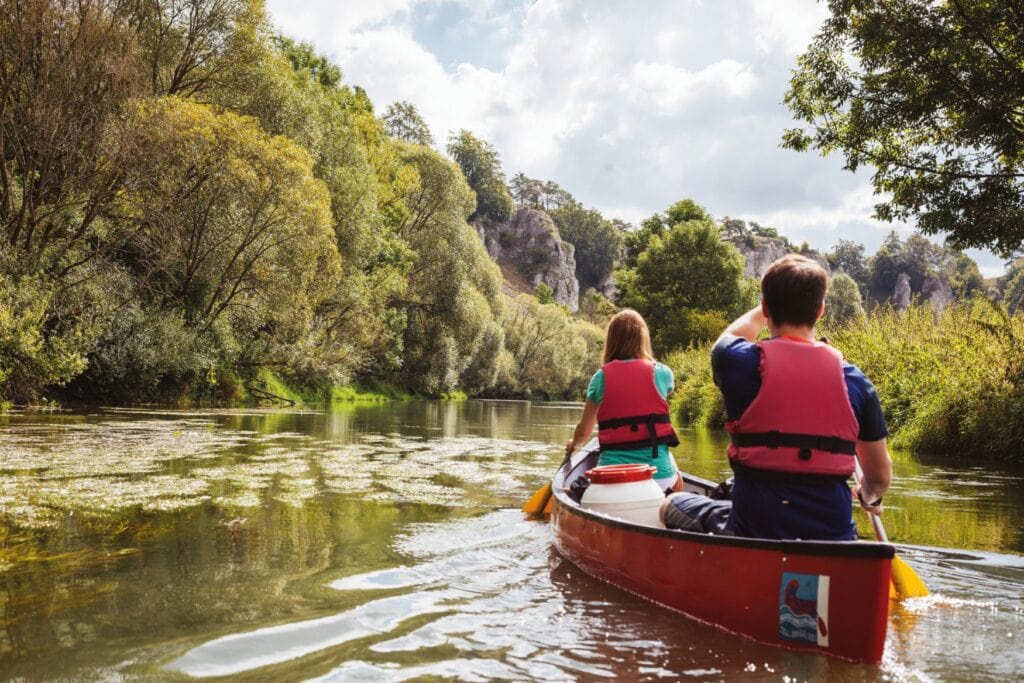 Zwei Personen mit Schwimmwesten fahren vom Betrachter weg mit einem Kanu auf einer nach rechts zeigenden Flussbiegung. Links am Ufer sind zunächst Wälder, aus denen im Hintergrund einige große Felsen hervorschauen. Das rechte Ufer ist von dichtem Gras bewachsen. Einige Wolken verdecken die Sonne.