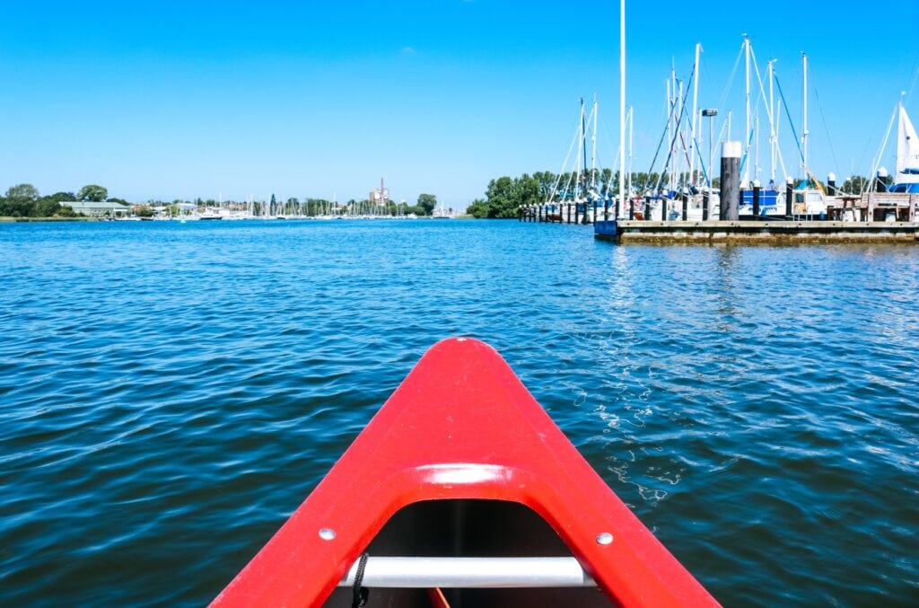 Die rote Spitze eines Kanus, vom Boot aus aufgenommen. Dahinter ein Haffen mit tiefblauem, ruhigen Wasser. Rechts und links reihen sich Boote mit ihren Masten aneinander, weit im Hintergrund sind einige Bäume am Ufer zu sehen