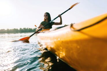 Eine Frau mit Sonnenbrille und grünem Shirt taucht ein Blatt ihres Doppelpaddels von einem knallgelben Kanu aus ins Wasser. Das Kanu fährt dabei rechts am Betrachter vorbei. Im Hintergrund ein bewaldetes Ufer.