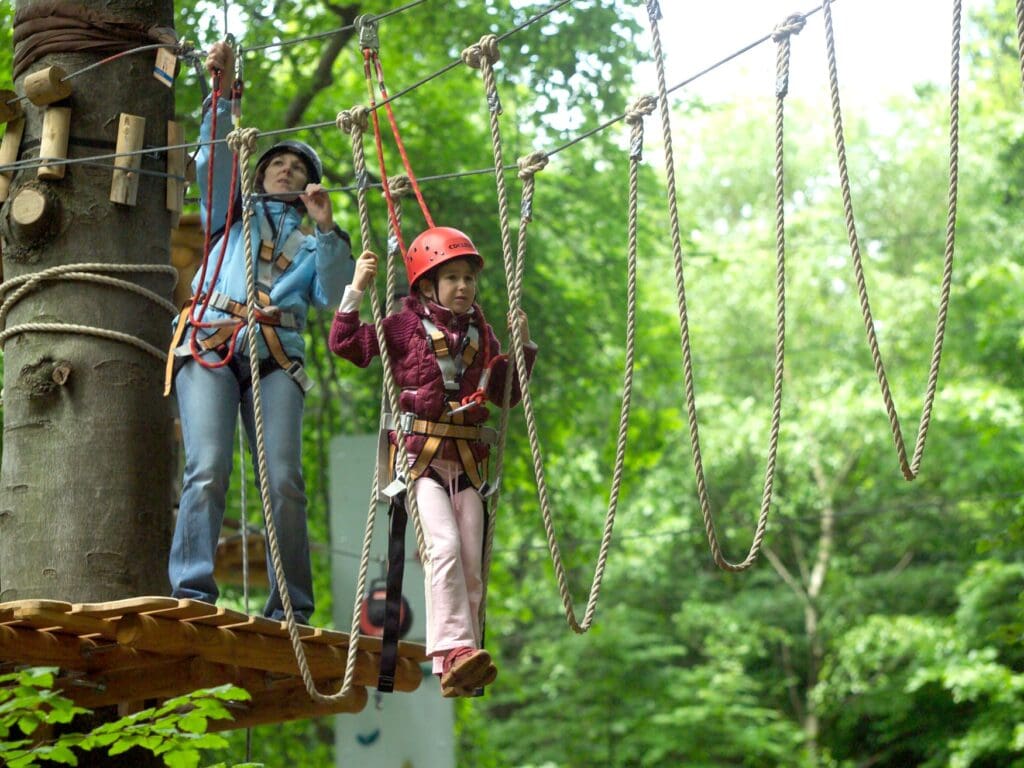Eine Frau und ein Kind mit Helm und Hüftgurt auf einem Baumwipfelpfad; das Kind steht mit beiden Beinen auf dem zweiten von mehreren Seilschlaufen, die Frau sichert sich gerade an der dahinter angebrachten Plattform.