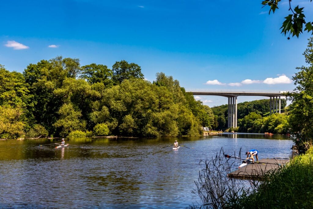 Eine Flussbiegung bei Sonnenschein. Am linken Ufer ist ein dichter Wald, am rechten Ufer macht gerade ein Mann sein Kanu an einem Anleger fest. Im Hintergrund spannt eine Autobahnbrücke über den Fluss.