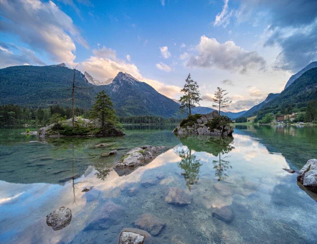 A clear blue lake with a few stone islands. On the other side of the lake the mountain ranges of the alps, nearly covered with forest, reach for the sky