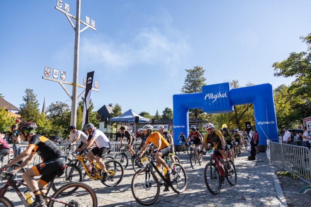 Zahlreicher Radler fahren auf einer durch Barrieren eingezäunten Strecke neben- und hintereinander von rechts nach links. An den Lenkern sind Startnummern befestigt. Hinter den Radlern spannt ein aufblasbarer, blauer Bogen mit der Aufschrift "Allgäu" über die Strecke. Über der Szene blauer Himmel